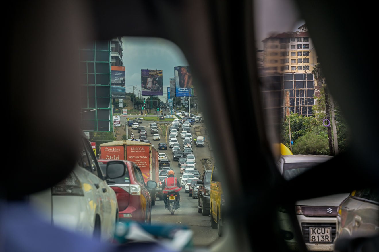 Kø av biler fyller en flerfelts vei i Nairobi, sett gjennom vinduene i et kjøretøy, mens en motorsykkel kjører mellom feltene. Bygninger, trafikklys og reklameskilt er synlige langs veien.