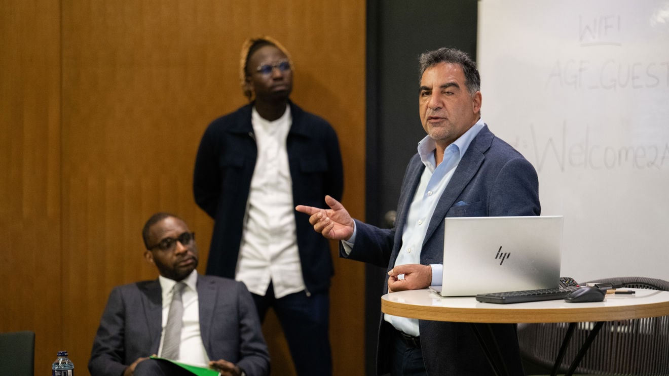 En mann i blå dress står ved en talerstol og snakker, mens han gestikulerer med hånden. To andre menn i dress følger med i et formelt konferanserom med trepanel på veggen og en stor skjerm ved siden av taleren.