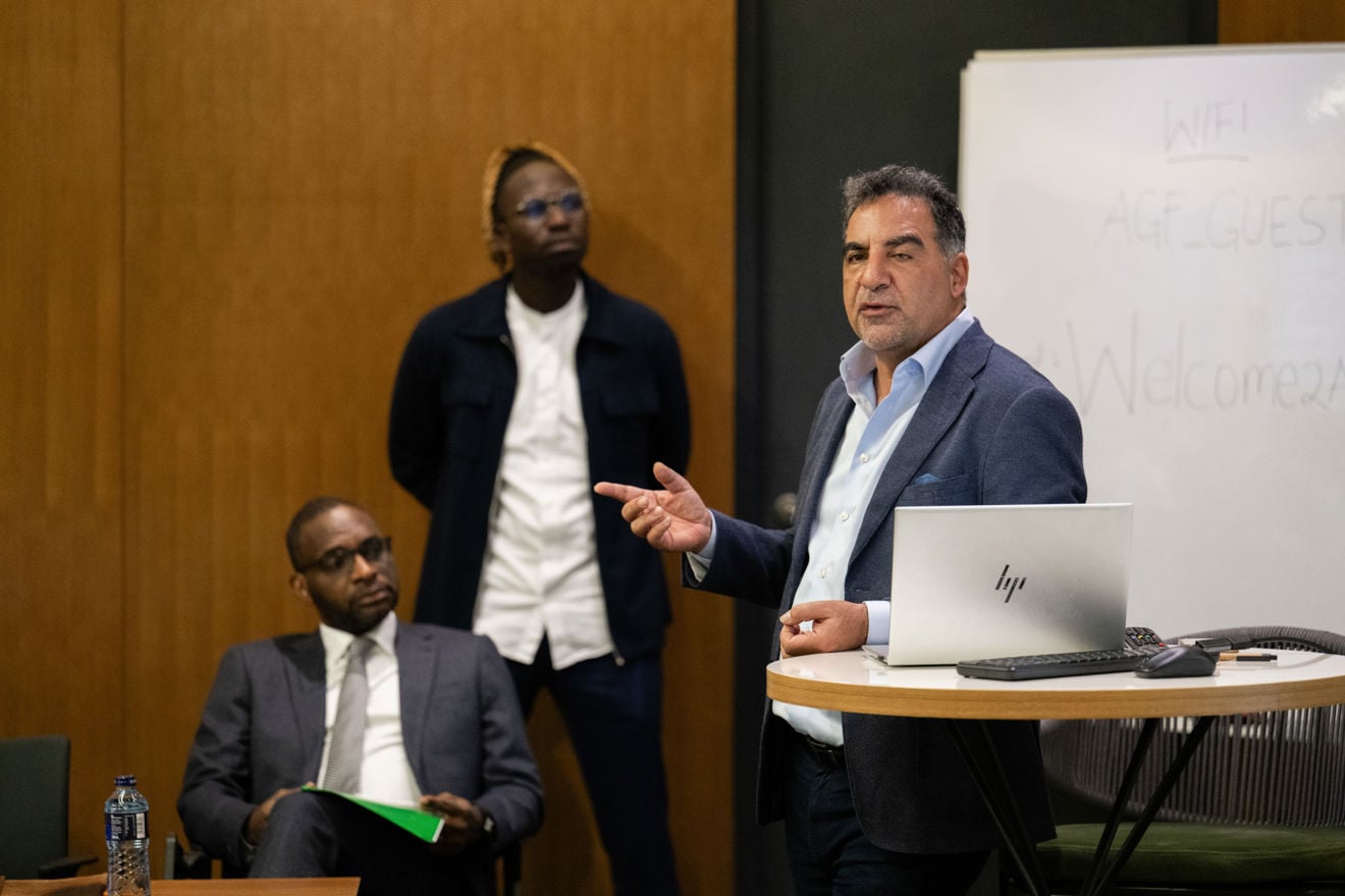 En mann i blå dress står ved en talerstol og snakker, mens han gestikulerer med hånden. To andre menn i dress følger med i et formelt konferanserom med trepanel på veggen og en stor skjerm ved siden av taleren.
