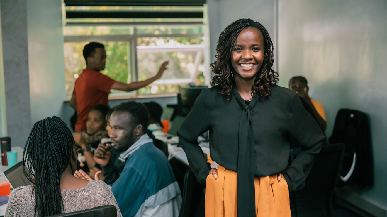 Winnie Ongiri står og smiler i et kontorlokale, mens flere personer sitter og arbeider ved datamaskiner i bakgrunnen