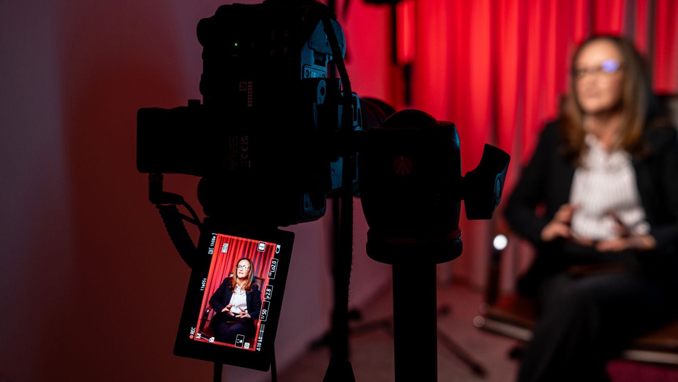 Bildet viser et filmkamera i forgrunnen som filmer en voksen kvinne i dress, sittende foran røde gardiner i et innendørs studio.