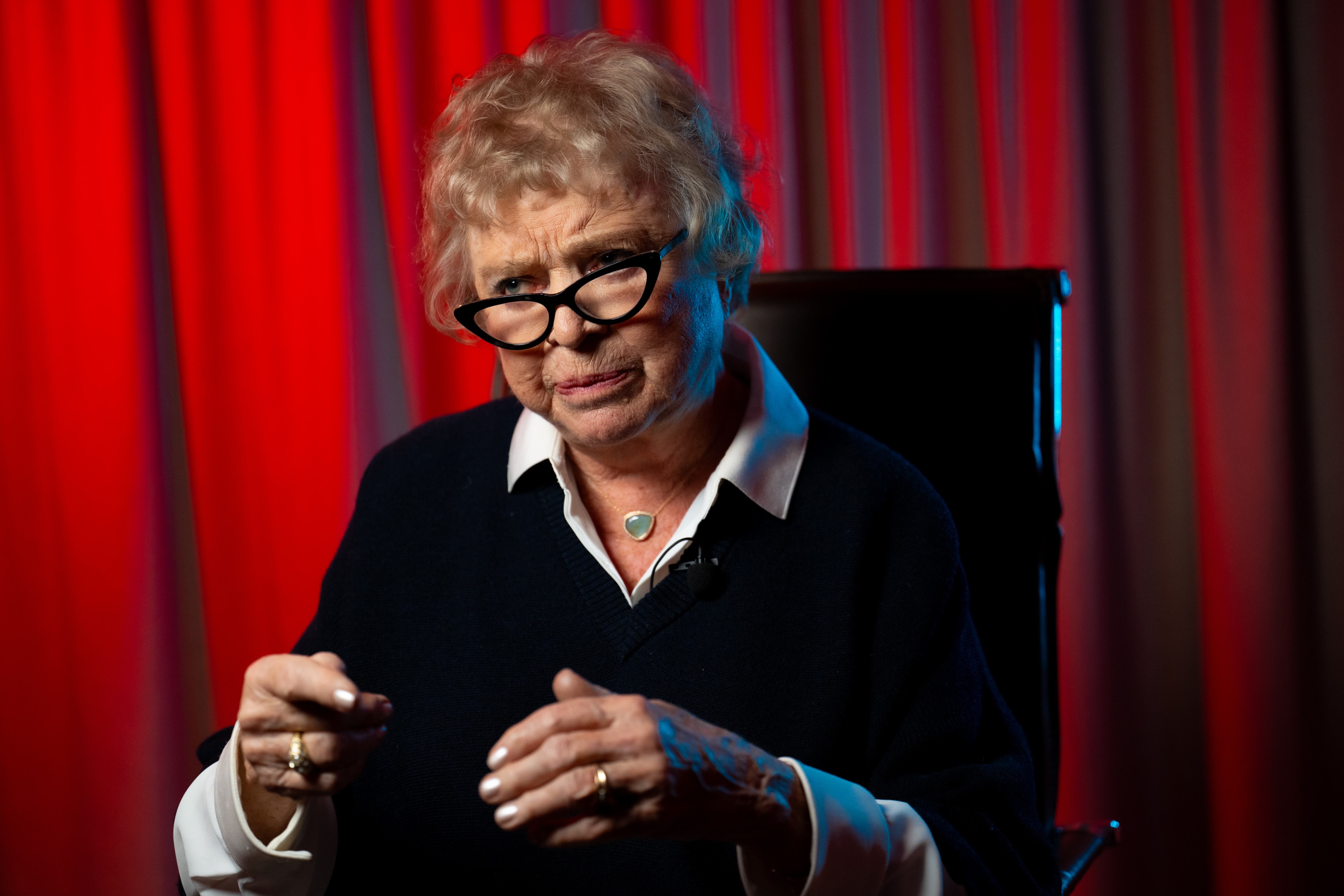 Bildet viser en eldre kvinne med lyst hår, mørk genser og hvit skjorte, sittende foran et bakteppe med røde gardiner og gestikulerende med hendene.