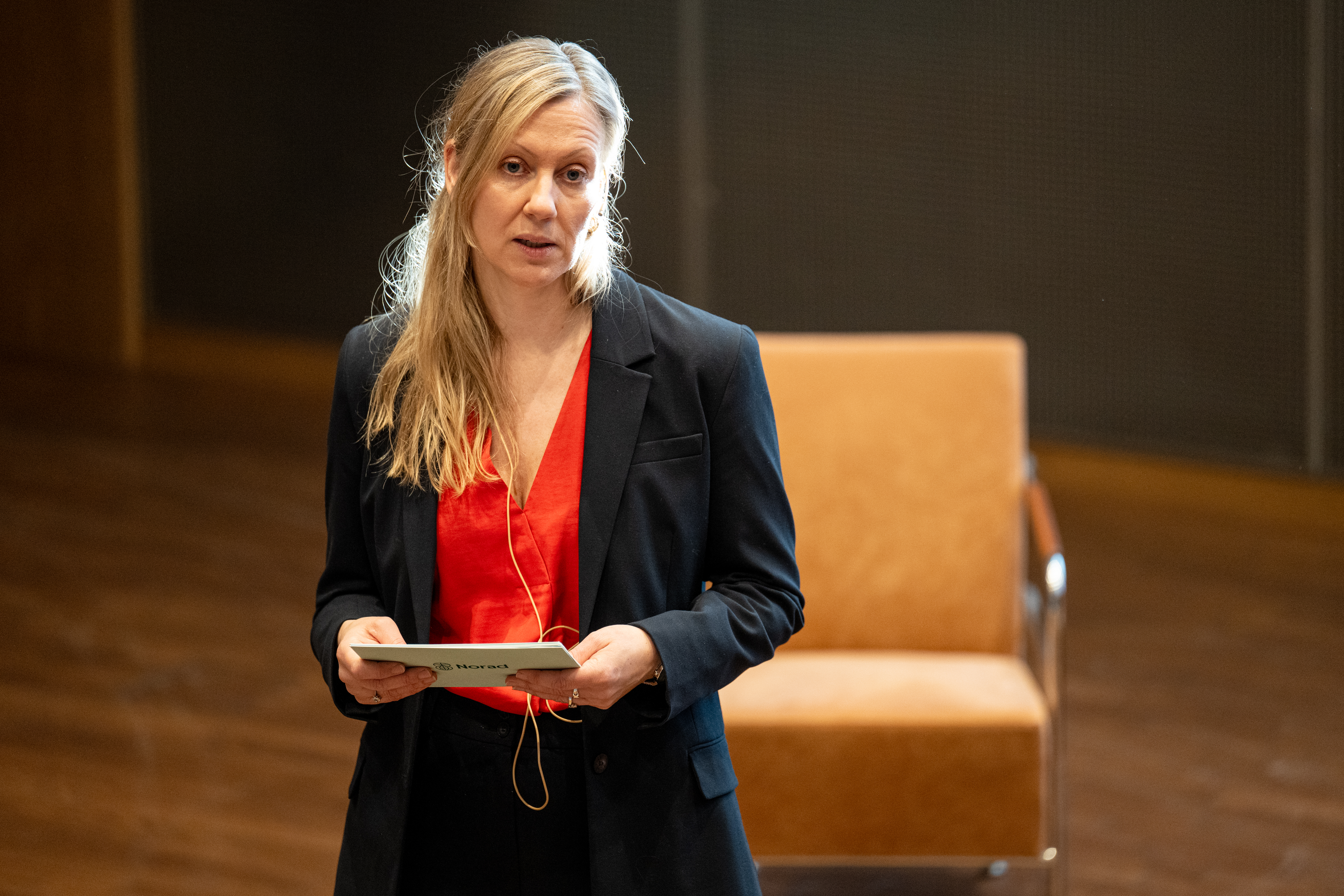 A young woman with long blonde hair, wearing a red blouse and dark blazer, stands indoors holding a notecard.