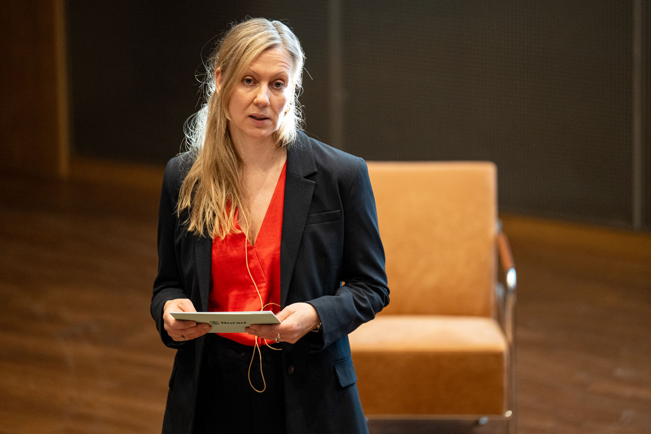 A young woman with long blonde hair, wearing a red blouse and dark blazer, stands indoors holding a notecard.