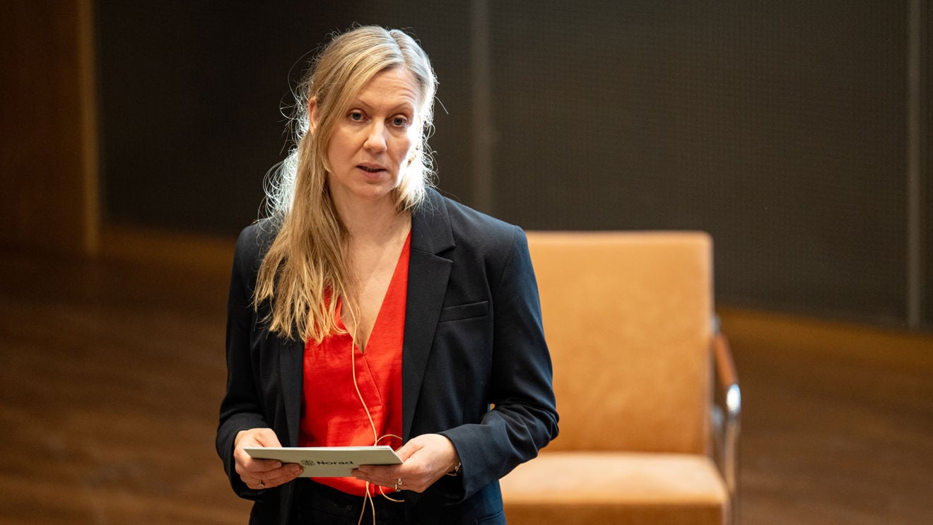 Bildet viser en ung kvinne med langt lyst hår, iført mørk dressjakke og rød topp, som står innendørs og holder et ark eller notatkort.