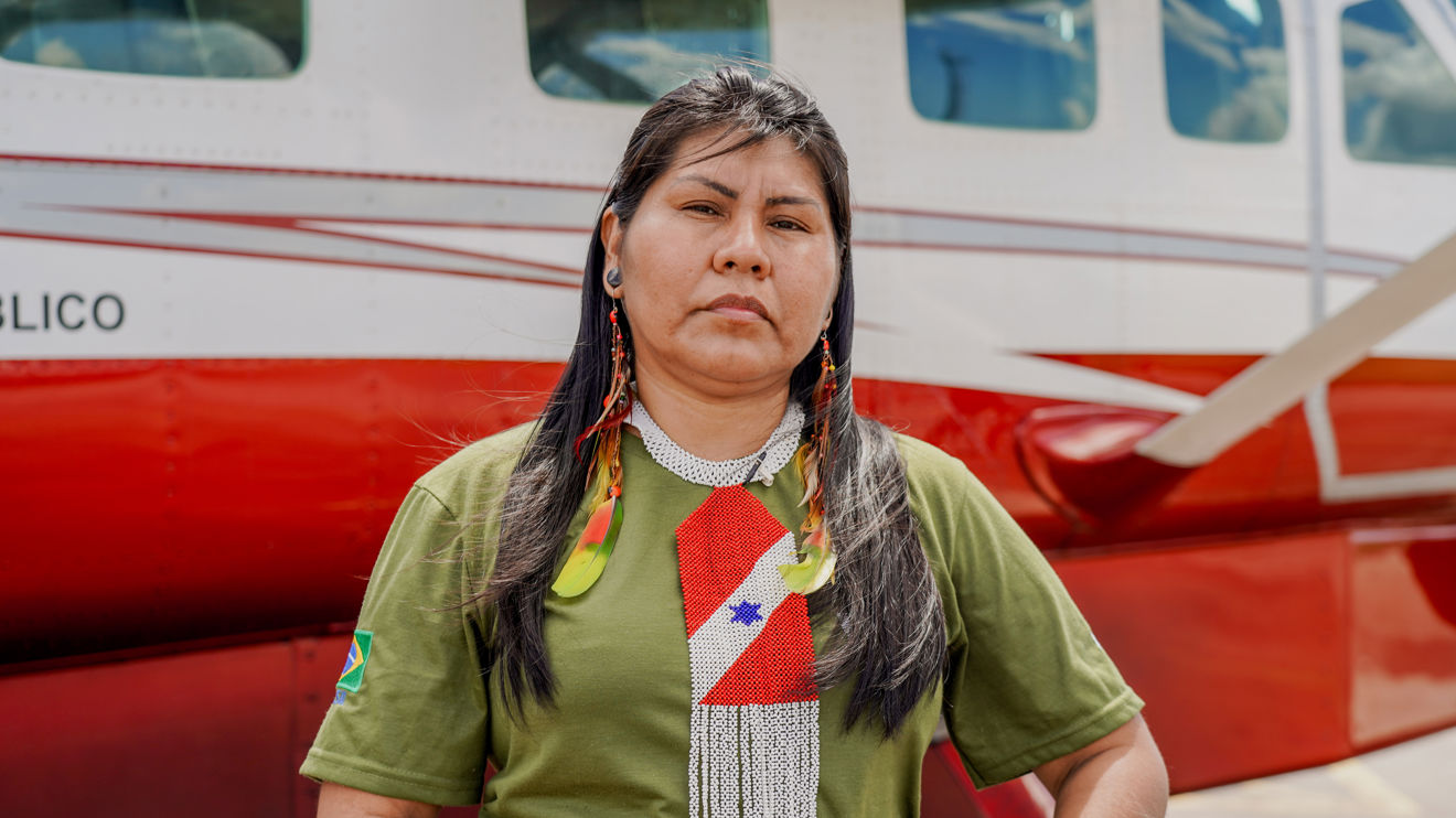 A woman with long dark hair, wearing a green shirt and beaded jewelry, stands in front of a red and white airplane.