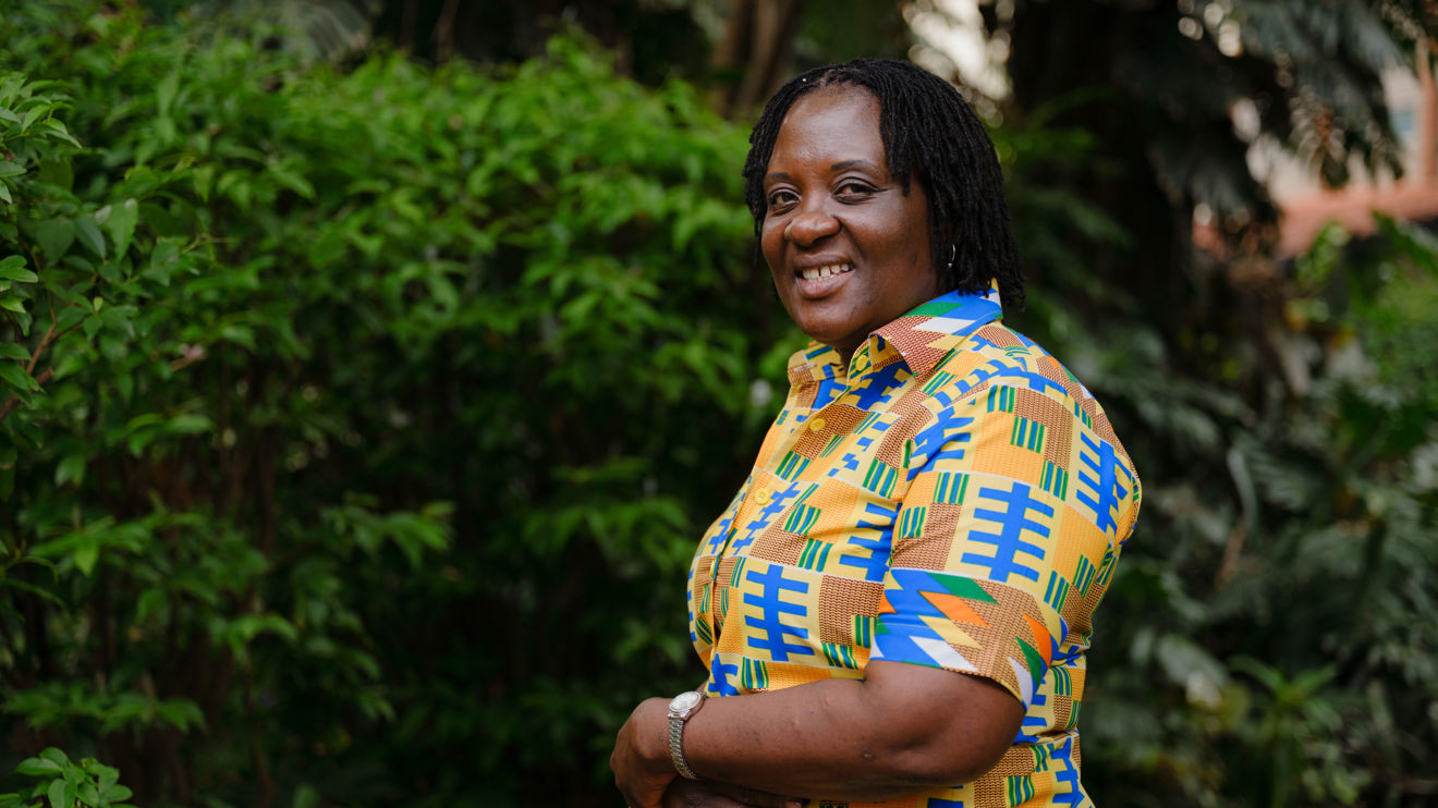 A woman wearing a colorful patterned shirt stands outdoors in front of green bushes.