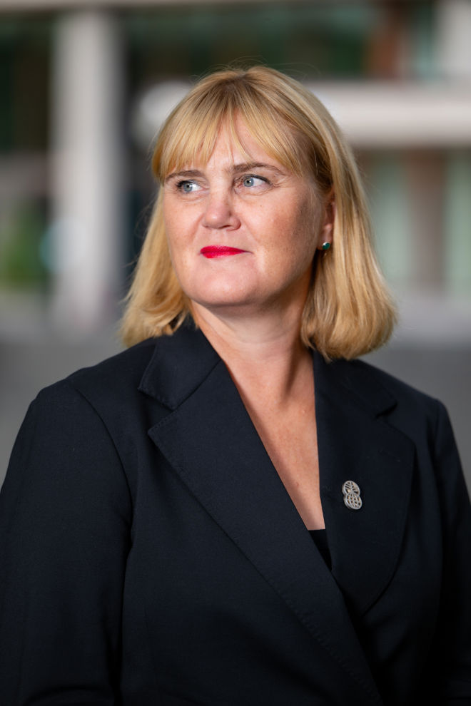 An adult woman with blonde hair wearing a dark blazer, photographed in an outdoor setting.