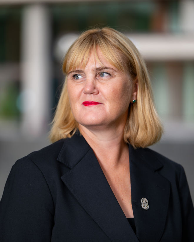 An adult woman with blonde hair wearing a dark blazer, photographed in an outdoor setting.