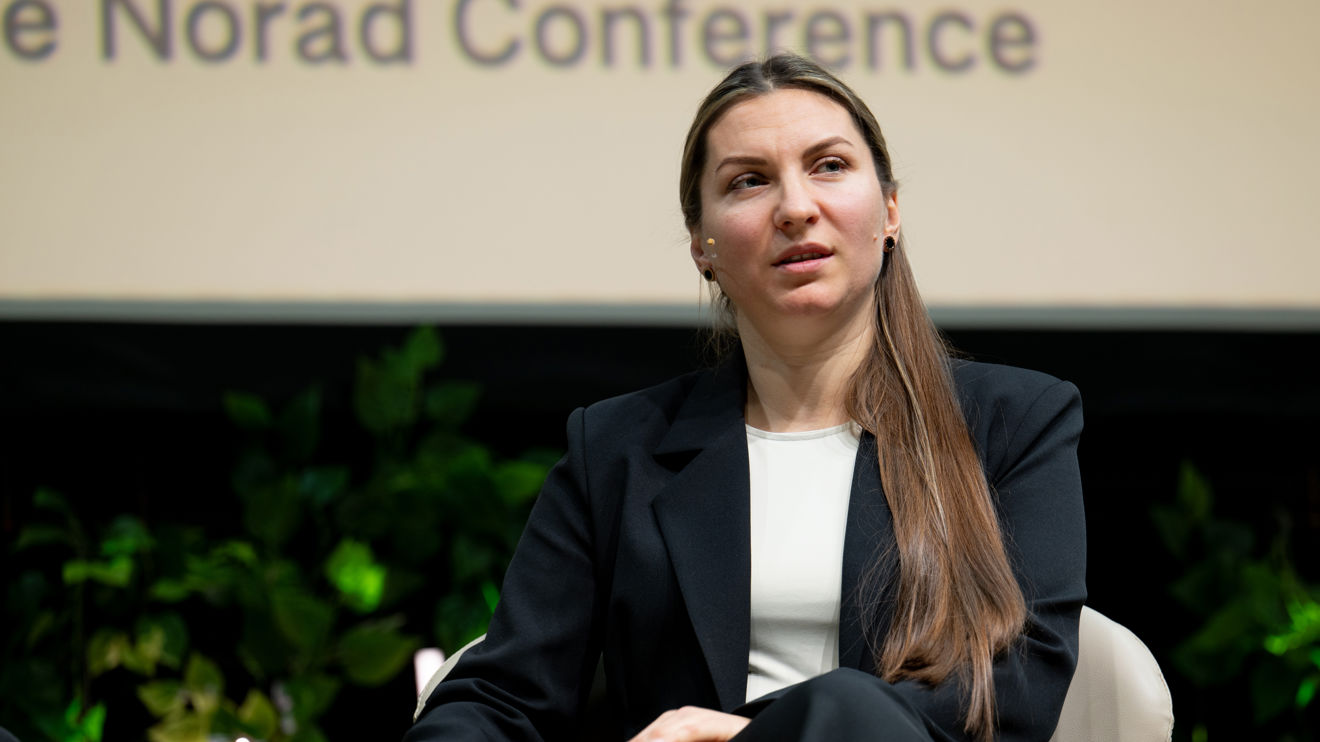 A woman with long brown hair, dressed in a black suit and white top, sits on a stage chair in front of a conference backdrop.