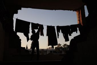 A woman hangs clothes on a line in a destroyed building, with the sky visible through large openings in the wall.