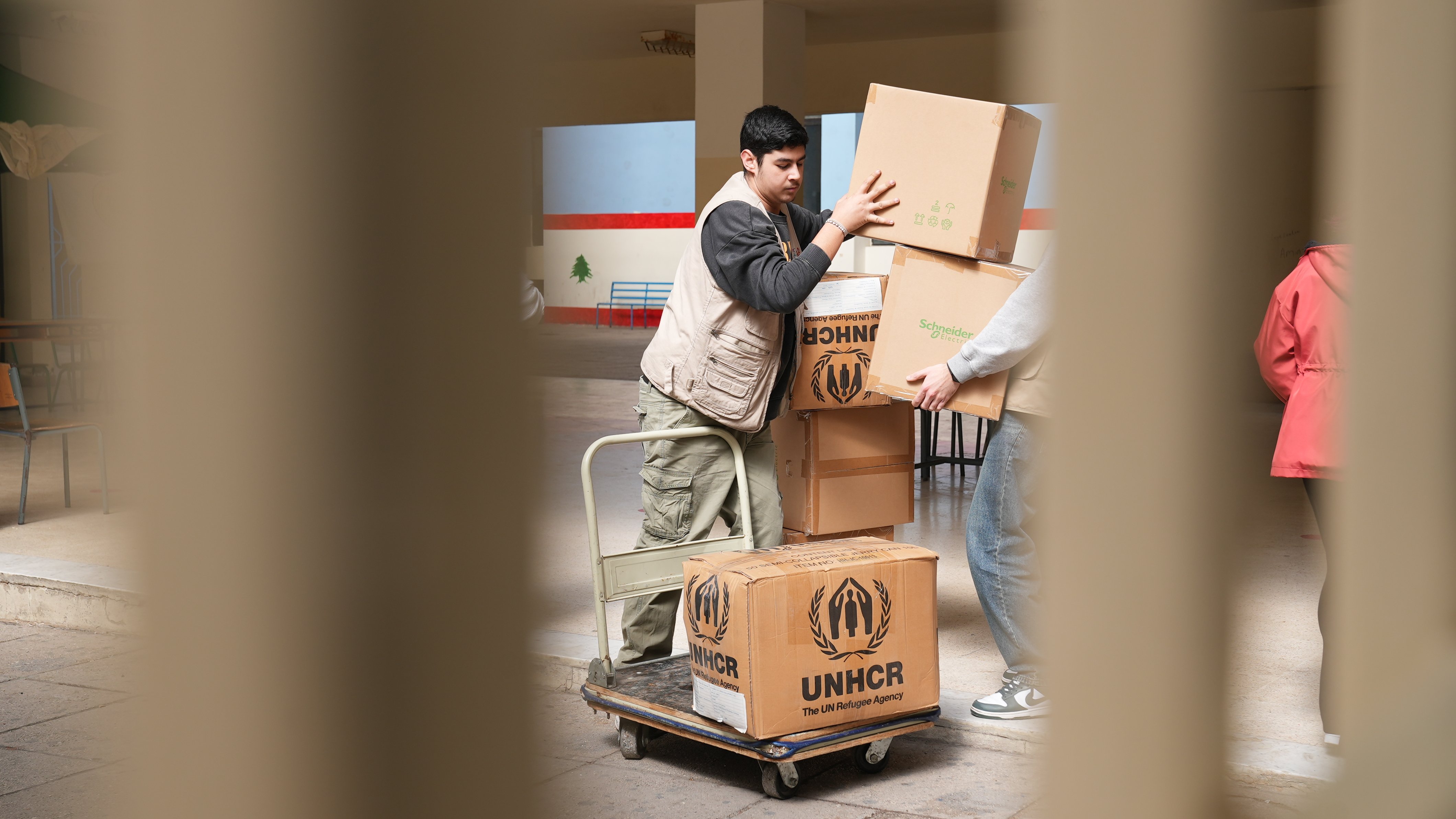 Bildet viser to unge menn innendørs som stabler esker merket med UNHCR-logo på en tralle.