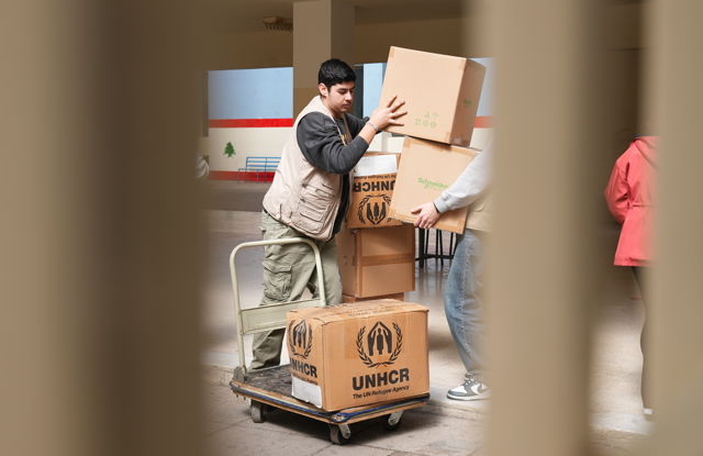 The image shows two young men indoors stacking boxes marked with the UNHCR logo onto a cart.