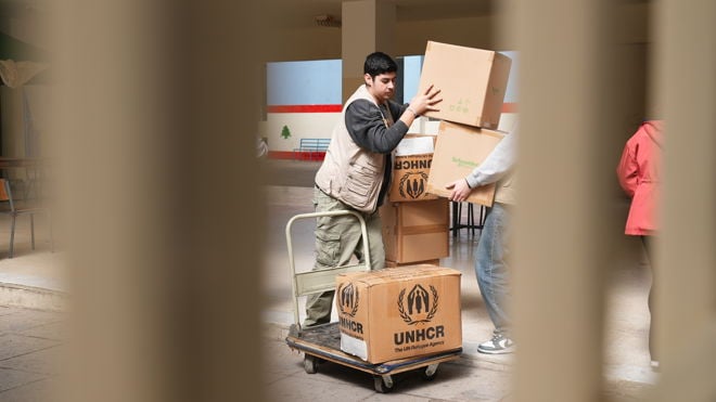 To voksne personer løfter pappesker med UNHCR-logo på en tralle innendørs.