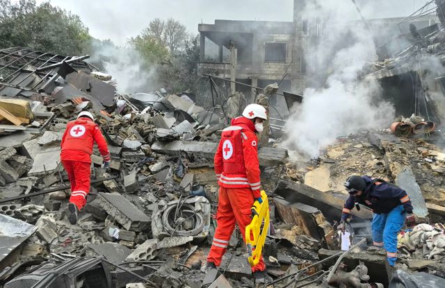 Redningsarbeidere i røde uniformer fra Røde Kors søker gjennom ruiner og røyken etter en ødelagt bygning.