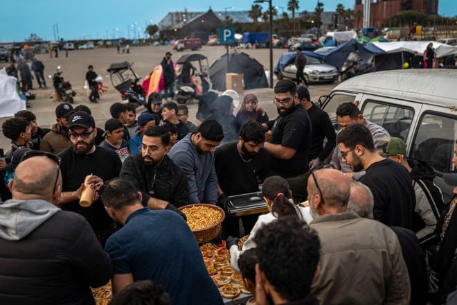 En stor gruppe voksne og unge samles rundt et bord utendørs for å få utdelt mat, med telt og kjøretøy i bakgrunnen.