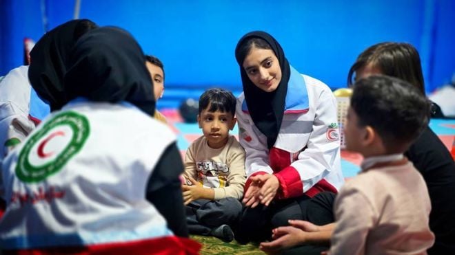 Flere voksne og barn sitter i ring på gulvet, hvor voksne i uniform fra Røde Halvmåne deltar i aktivitet med barna.