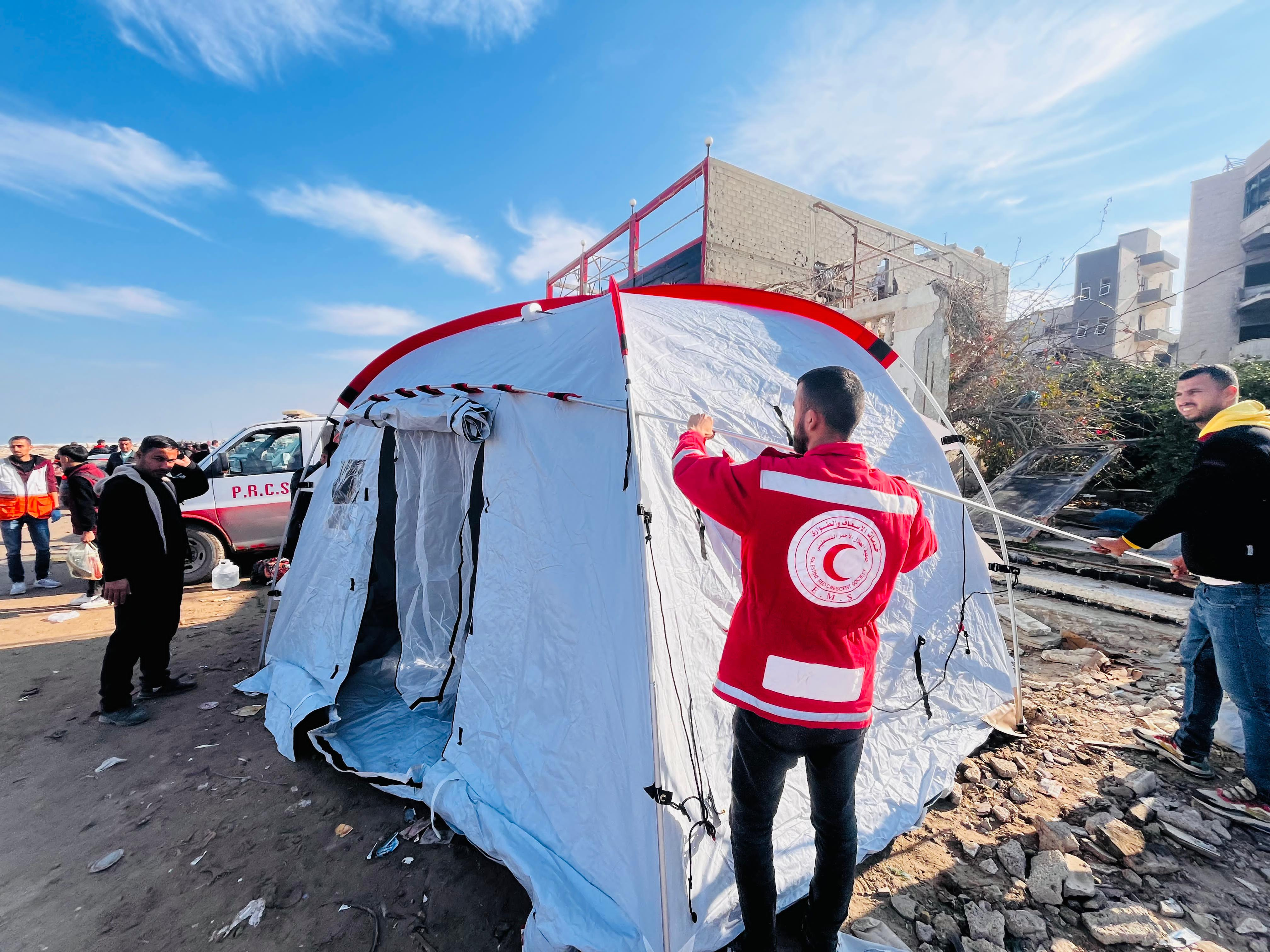 Flere voksne menn setter opp et hvitt telt med Røde Halvmåne-logo i et utendørs område med ødelagte bygninger i bakgrunnen.