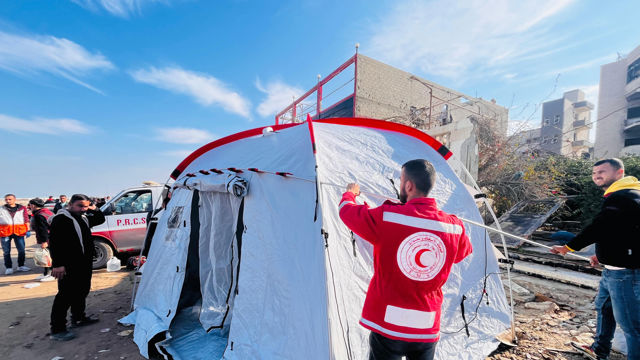 Flere voksne menn setter opp et hvitt telt med Røde Halvmåne-logo i et utendørs område med ødelagte bygninger i bakgrunnen.