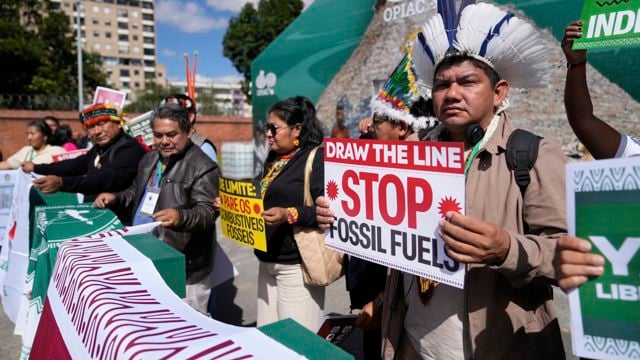 En gruppe demonstranter står samlet utendørs foran en bygning og holder plakater og grønne flagg. En mann i forgrunnen holder en plakat med teksten «Stop funding fossil fuels», mens flere personer rundt ham roper eller holder opp slagord.