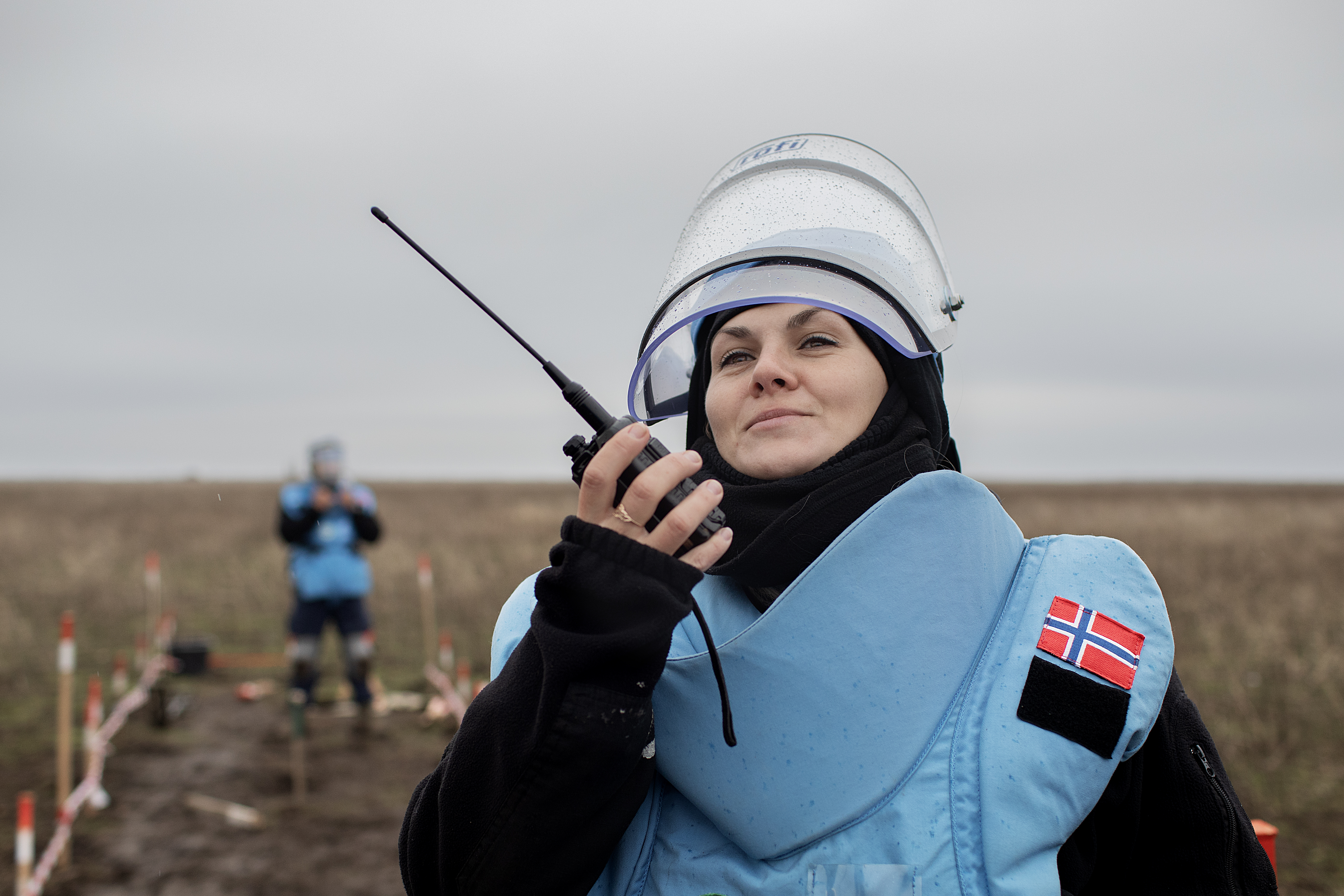 En person med beskyttelsesvest, visir og norsk flagg på skulderen holder en walkie-talkie på et avsperret felt utendørs.