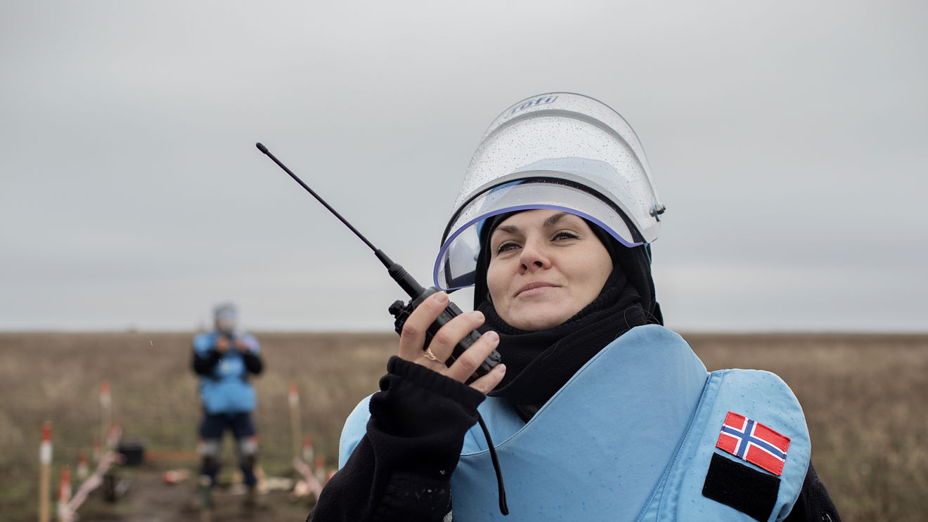 En person med beskyttelsesvest, visir og norsk flagg på skulderen holder en walkie-talkie på et avsperret felt utendørs.
