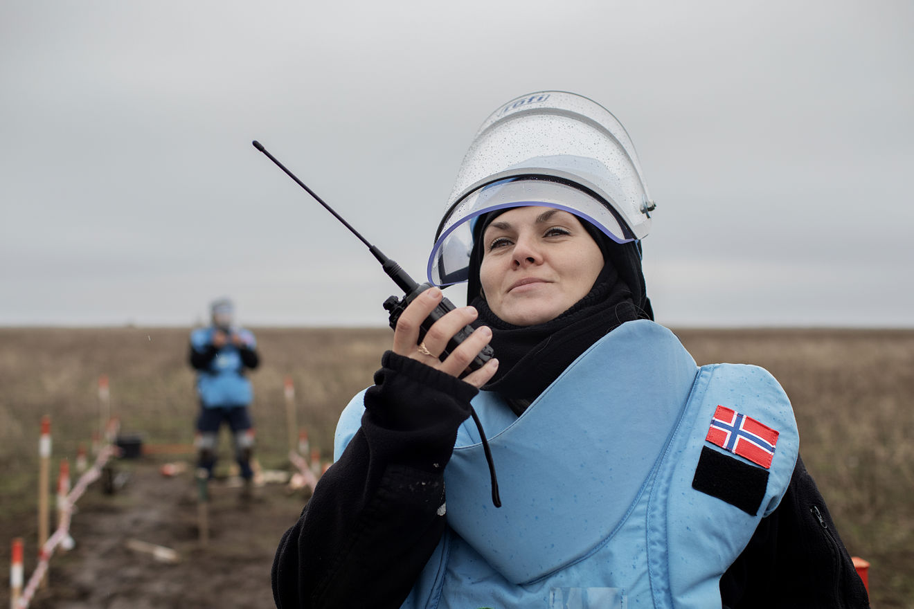 En person med beskyttelsesvest, visir og norsk flagg på skulderen holder en walkie-talkie på et avsperret felt utendørs.
