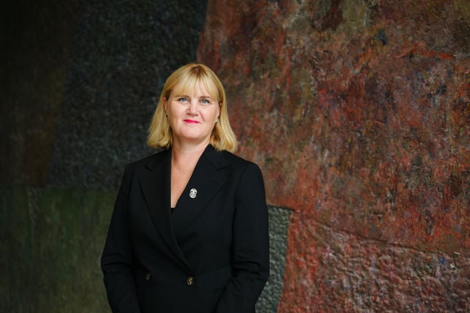 En kvinne med blondt hår og sort dressjakke står foran en vegg med rødt og grønt mønster.