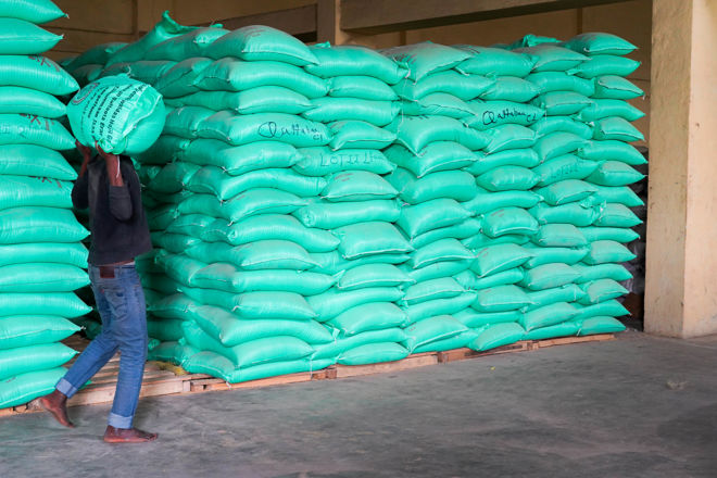 En person bærer en stor sekk foran en stable med grønne sekker inne i et lager.