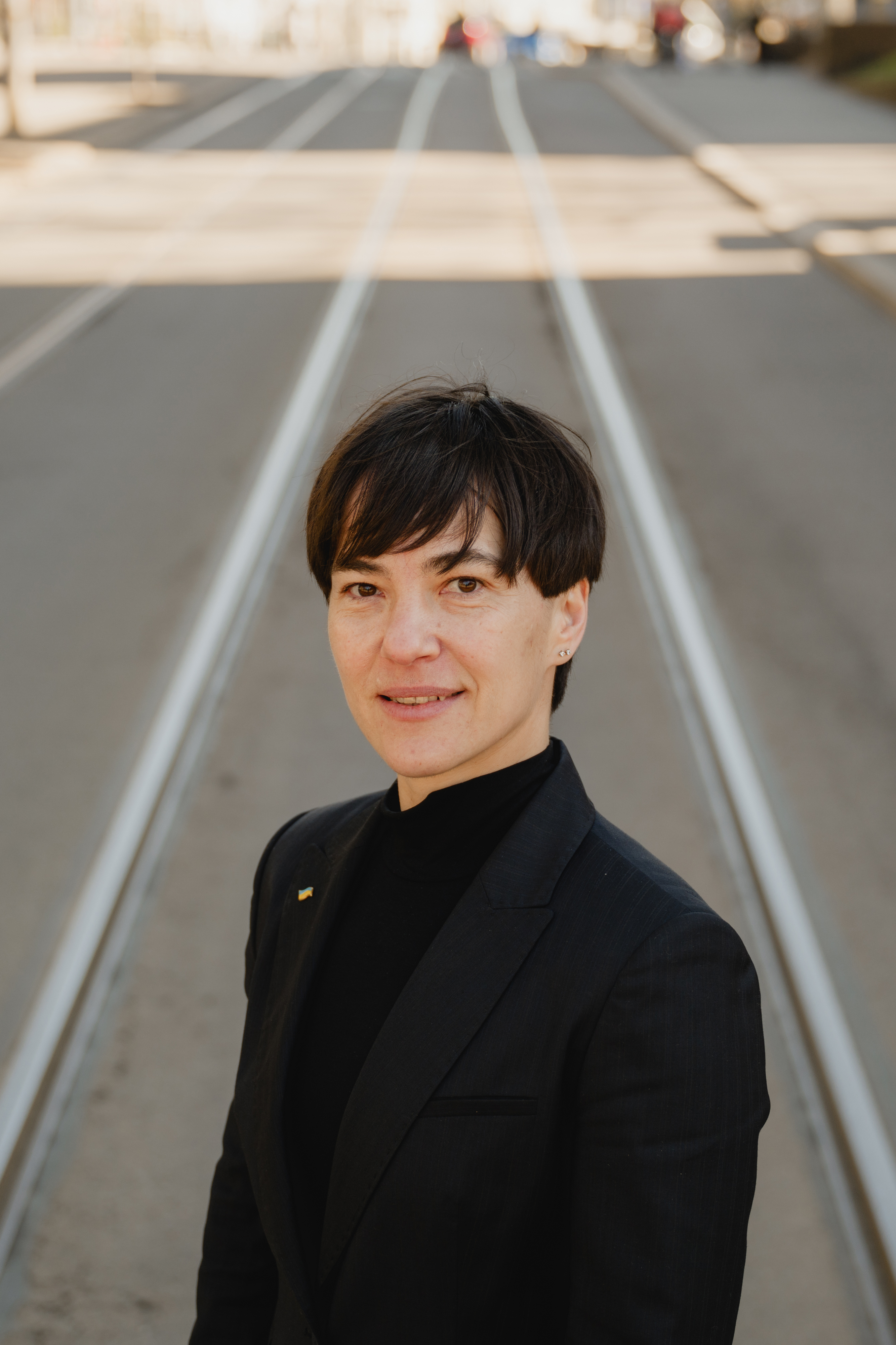 A young person stands on tram tracks in an empty street, wearing a dark jacket and black top.