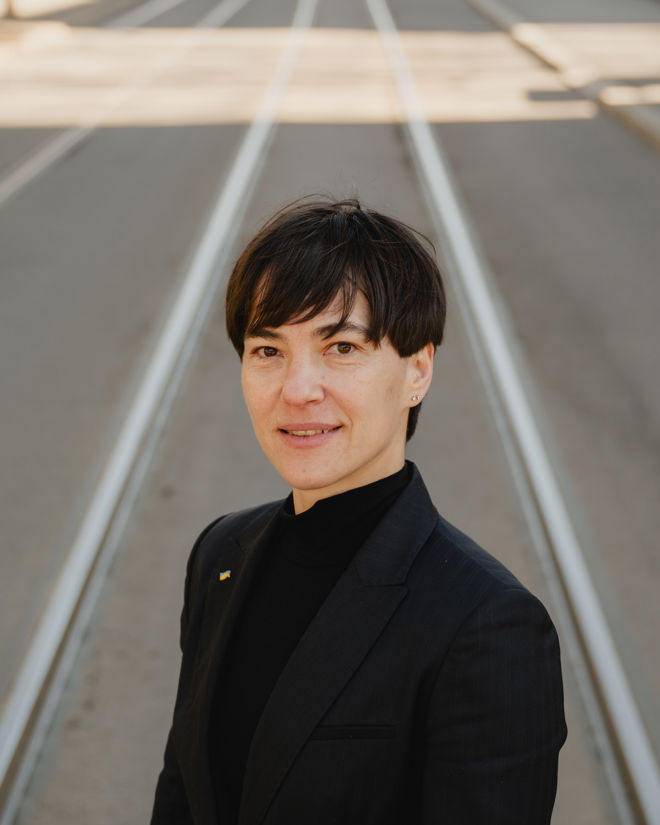 A young person stands on tram tracks in an empty street, wearing a dark jacket and black top.