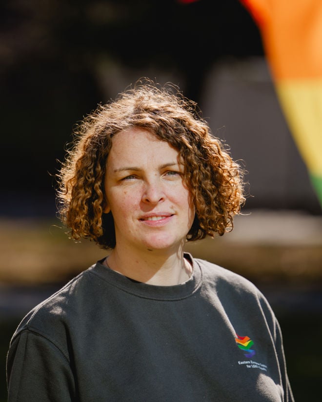A young person stands outdoors in front of a rainbow flag, wearing a grey sweatshirt.