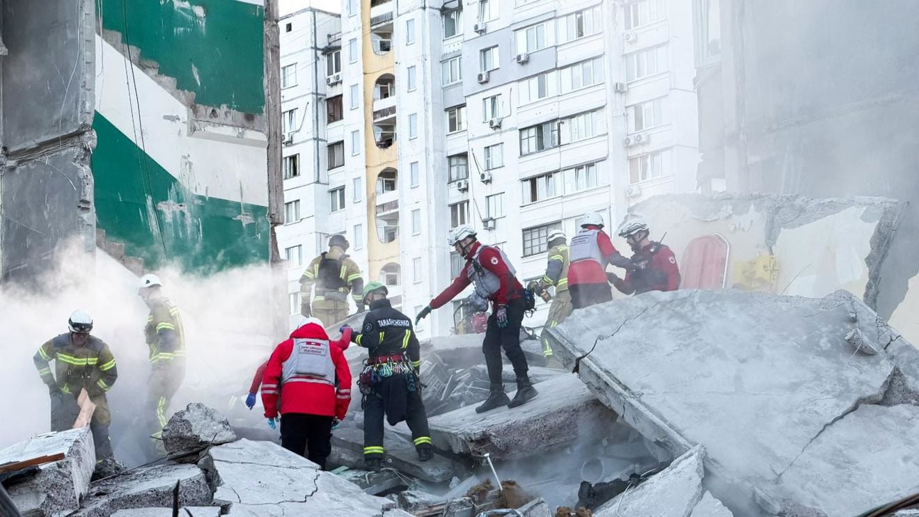 Redningsarbeidere med hjelmer og uniformer søker gjennom ruiner og sammenraste betongblokker etter et bygningsras i et urbant område.