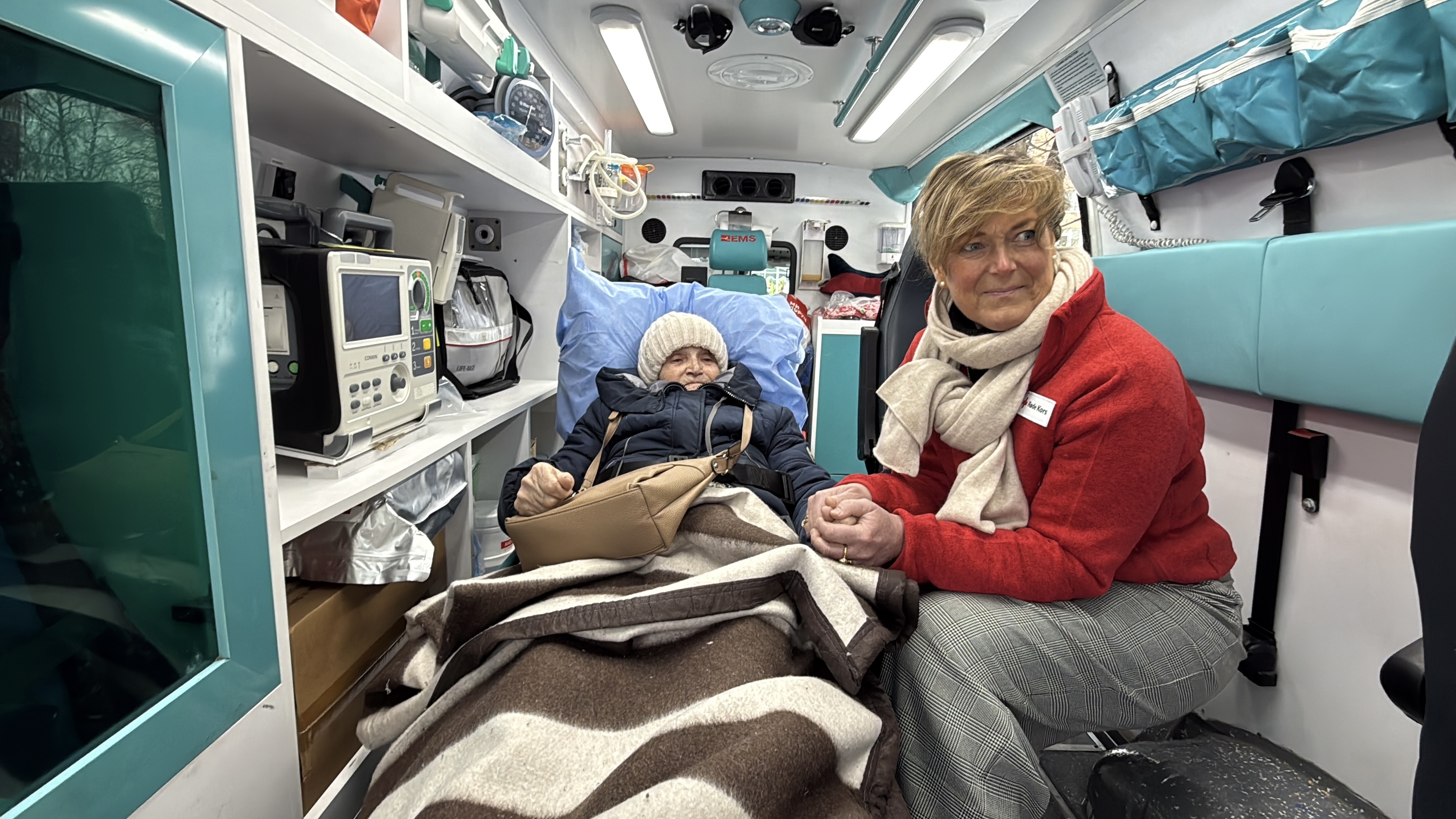 En eldre person ligger under et teppe på båre inne i en ambulanse, mens en voksen kvinne i rød jakke sitter ved siden av og holder personen i hånden.