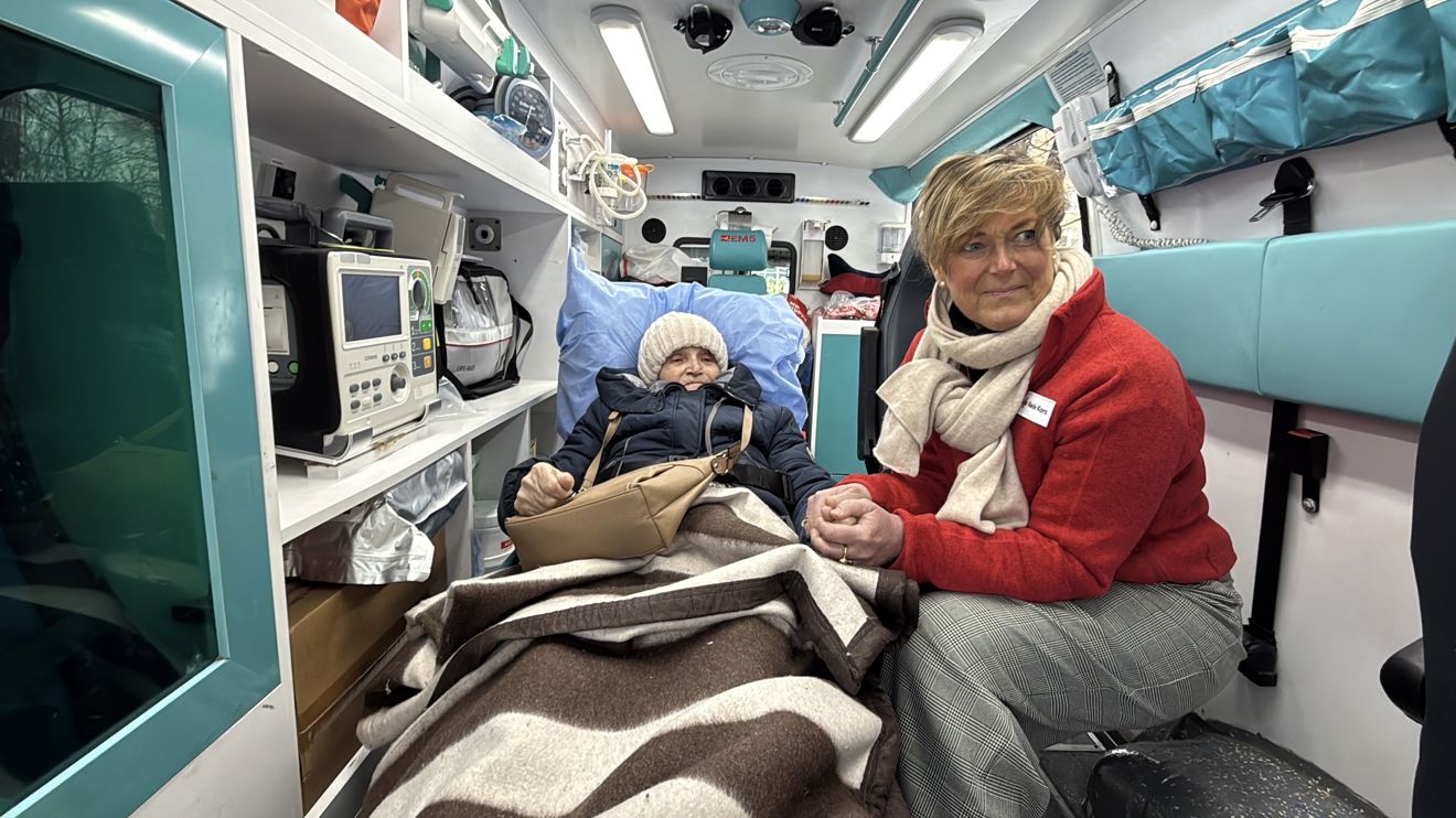 En eldre person ligger under et teppe på båre inne i en ambulanse, mens en voksen kvinne i rød jakke sitter ved siden av og holder personen i hånden.