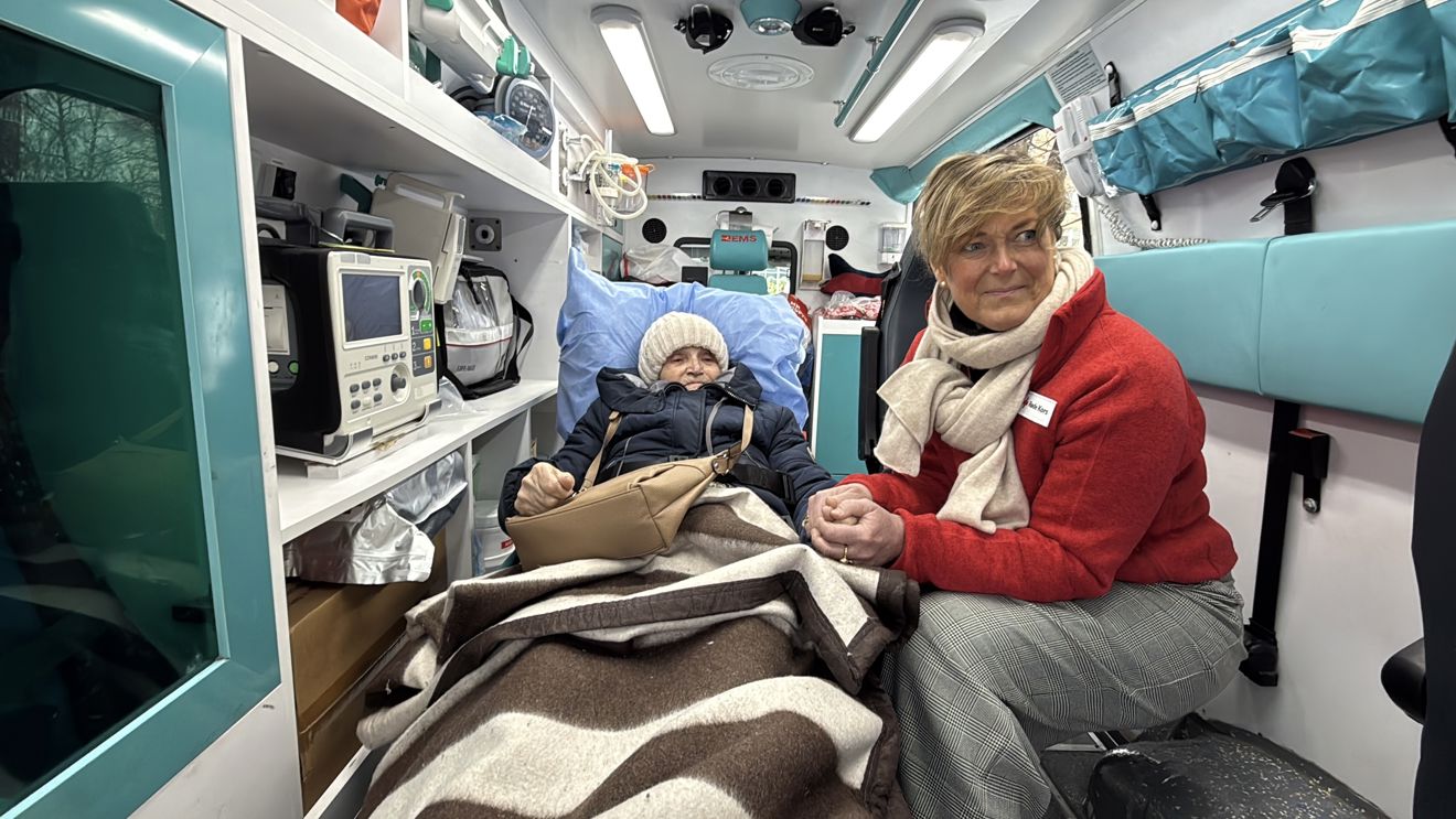 En eldre person ligger under et teppe på båre inne i en ambulanse, mens en voksen kvinne i rød jakke sitter ved siden av og holder personen i hånden.