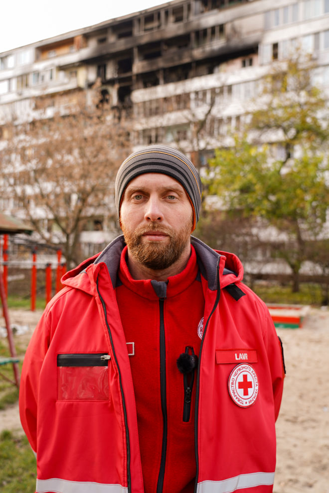 En voksen person med rød Røde Kors-jakke og lue står foran en boligblokk med brannskader på fasaden.