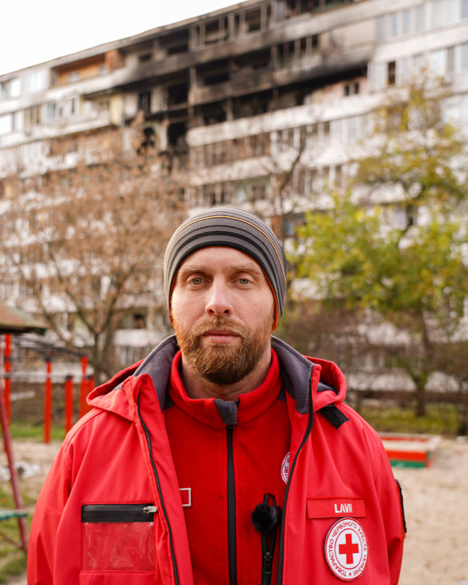 En voksen person med rød Røde Kors-jakke og lue står foran en boligblokk med brannskader på fasaden.