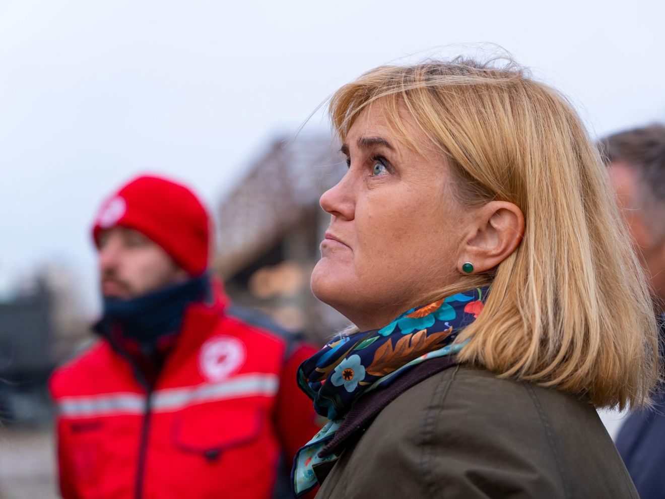 En voksen kvinne med lyst hår og skjerf står utendørs sammen med en person i rød Røde Kors-uniform.