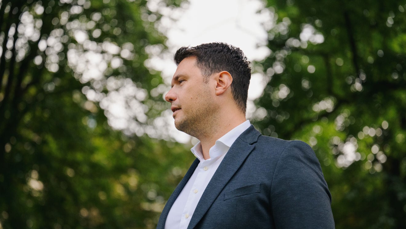 En mann i mørk blazer og hvit skjorte står utendørs, fotografert fra siden.