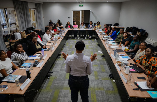 A woman stands in front of a group of participants seated around a U-shaped conference table and speaks in a meeting room. The participants have notebooks, bottles, and name cards in front of them.