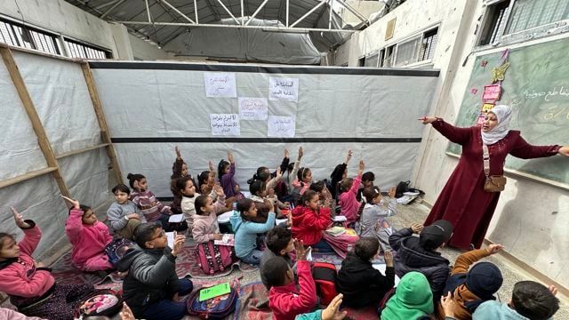 Lærer underviser en gruppe barn som sitter på gulvet i et provisorisk klasserom, mens flere av barna rekker opp hånden. Tavle, skolebøker og sekker er synlige i rommet.