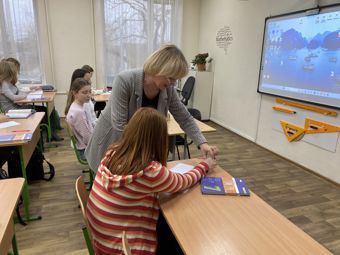 En lærer hjelper en elev ved pulten i et klasserom, mens flere andre elever sitter og jobber.