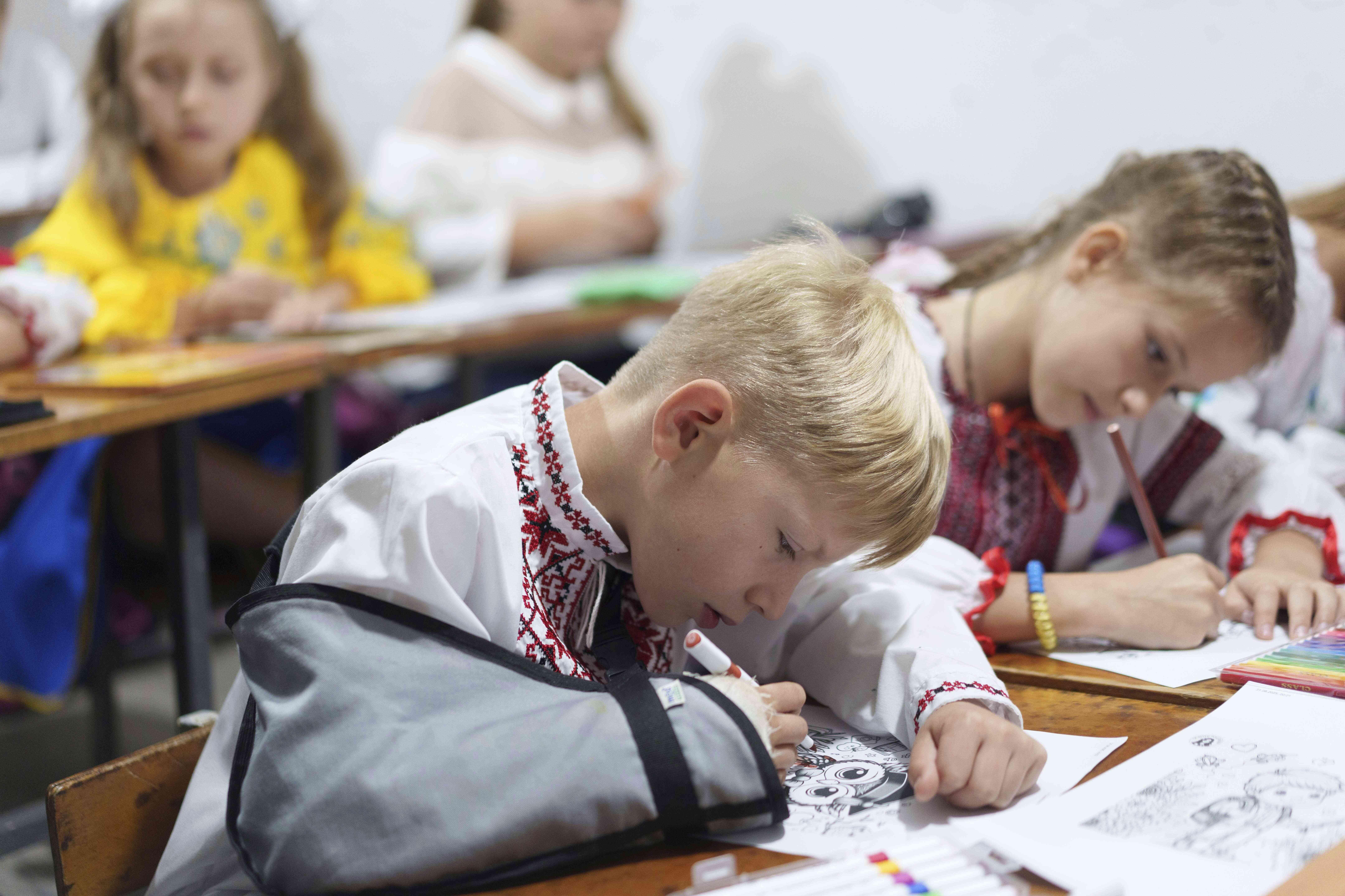 Barn kledd i tradisjonelle klær sitter ved pulter og fargelegger tegninger.
