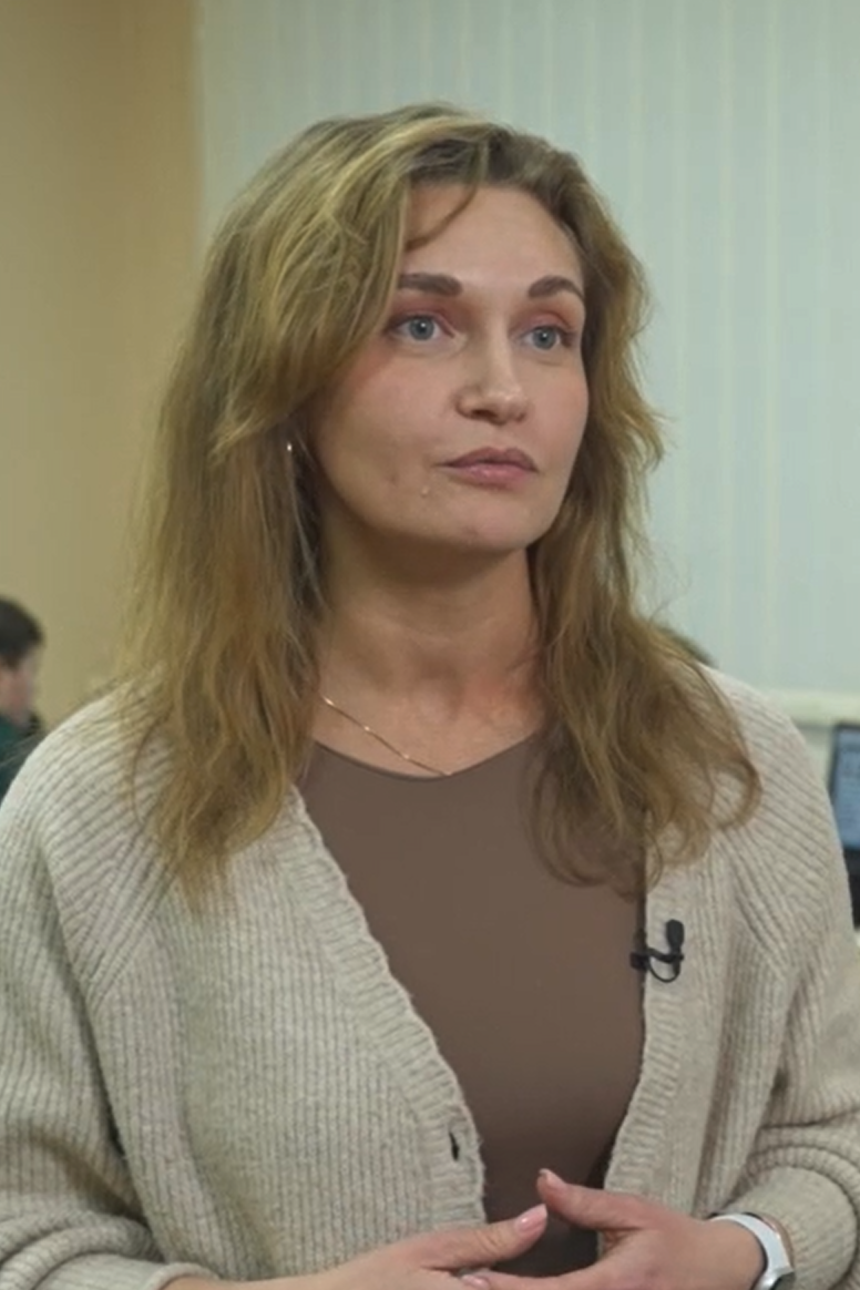 En voksen kvinne står innendørs og snakker, iført beige cardigan og mikrofon festet på brystet.