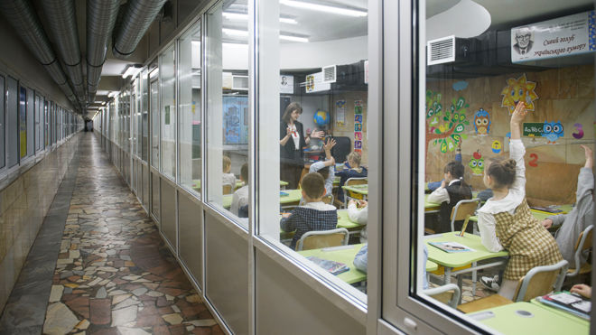 Barn sitter i et klasserom med fargerike veggdekorasjoner, mens en lærer underviser og flere elever rekker opp hånden; klasserommet ligger i en korridor med glassvegger.