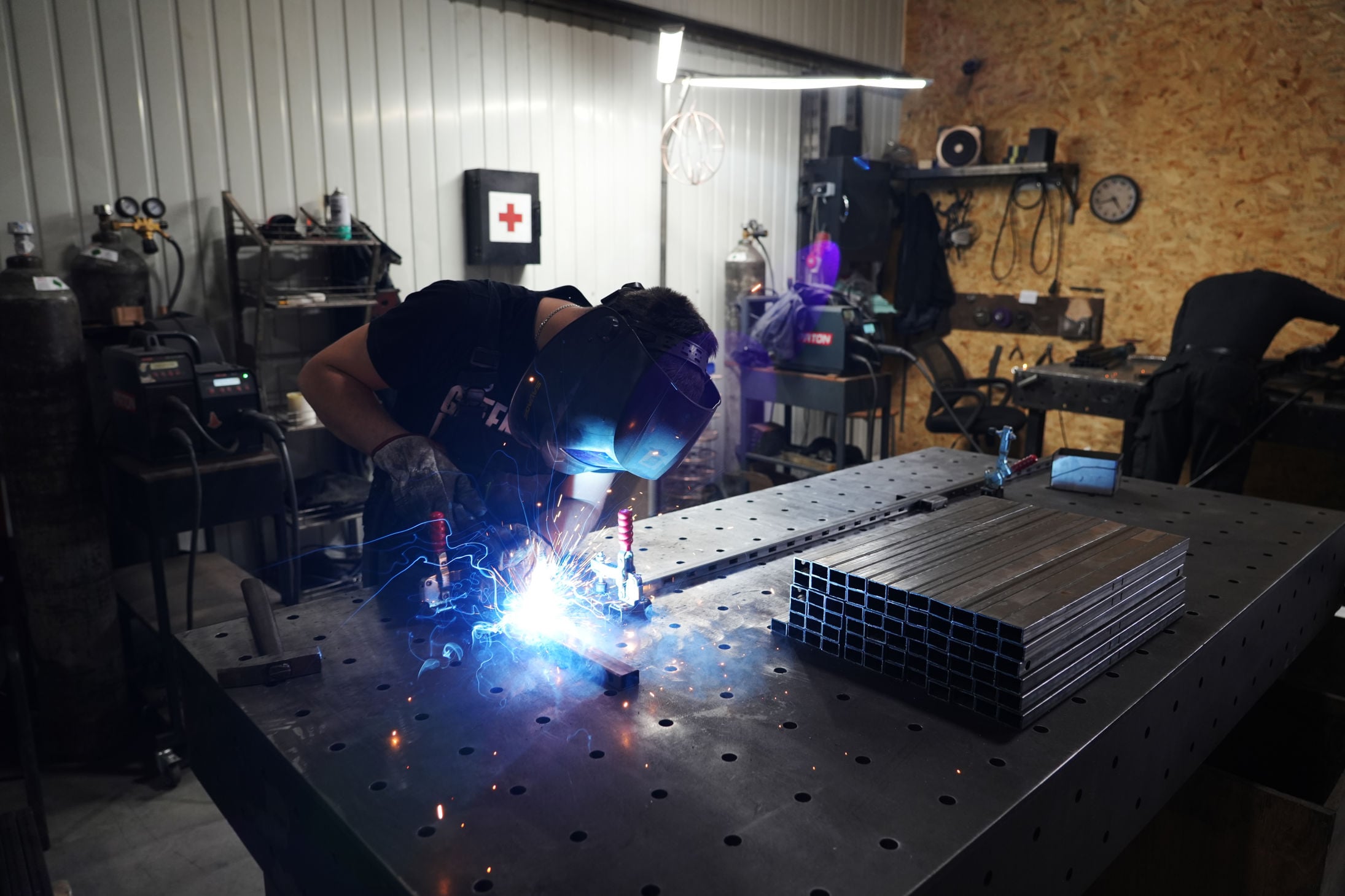 A person wearing a welding helmet is welding metal pieces on a worktable in a workshop. A stack of square metal tubes is placed on the table beside them.