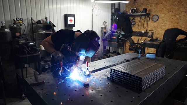 A person wearing a welding helmet is welding metal pieces on a worktable in a workshop. A stack of square metal tubes is placed on the table beside them.