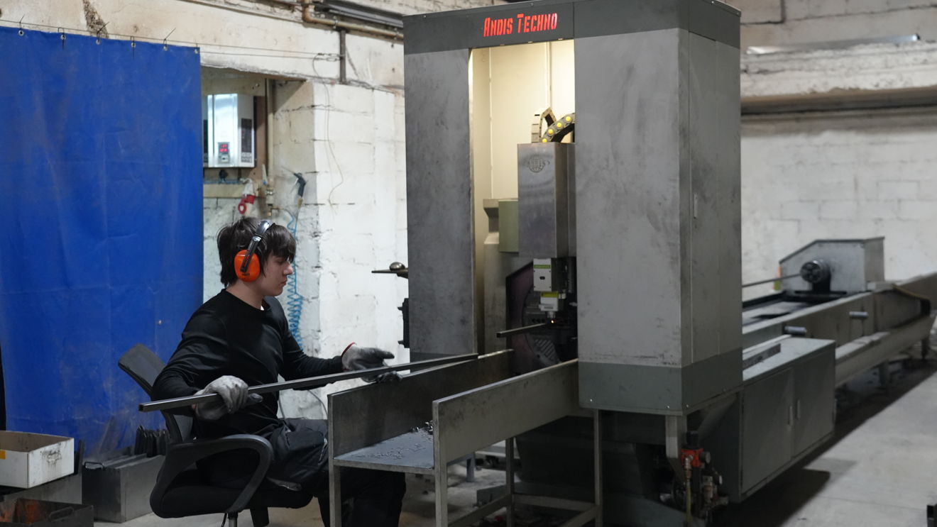 A person wearing ear defenders and gloves operates a metalworking machine in an industrial workshop. They are seated beside the machine and guiding a long metal bar into it.