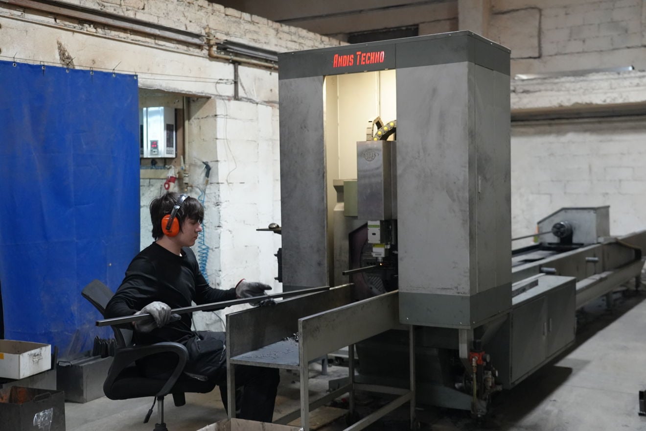A person wearing ear defenders and gloves operates a metalworking machine in an industrial workshop. They are seated beside the machine and guiding a long metal bar into it.